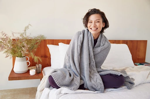 Cheerful woman wrapped in gray cable-knit blanket sitting cross-legged on a white bed in a brightly lit bedroom with natural light.