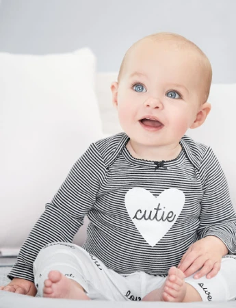 Adorable baby with bright blue eyes smiles while wearing a striped shirt with 'cutie' printed on a heart, sitting against a white background.