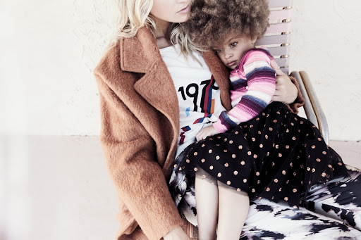 A woman in a coat sits holding a young child with curly hair in her lap. Both are posed in front of a neutral background.