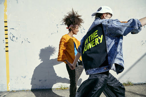 Two people walking outdoors, one with curly hair in orange top, the other in a denim jacket, with graffiti wall and shadows.