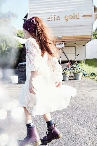 A young girl in a white dress and red boots walks past an off-white camper with a red headscarf on a sunny day in an outdoor setting.