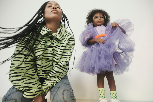 A young woman wearing a green and black tiger-striped jacket and jeans, and a young girl in a lilac dress are posed in a studio.
