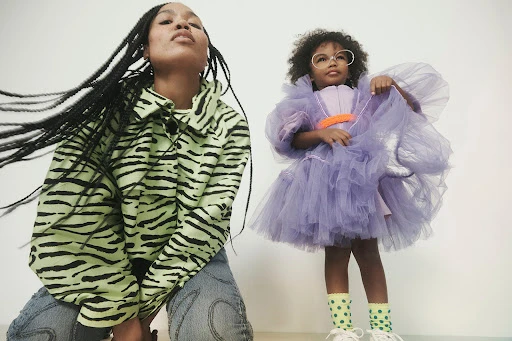 A young woman wearing a green and black tiger-striped jacket and jeans, and a young girl in a lilac dress are posed in a studio.