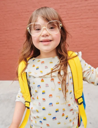 Adorable young girl with Down syndrome smiles in clear glasses and a yellow backpack, standing against a red brick background.