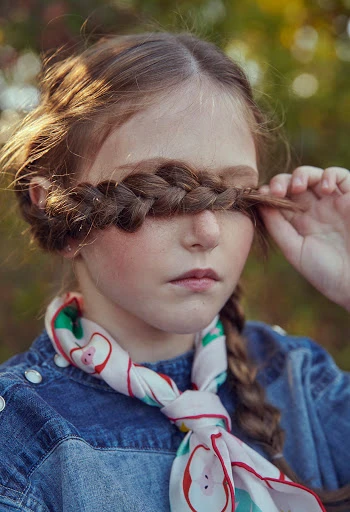 Young girl portrait with braided hair covering eyes, denim top, and apple-patterned scarf, posing in a softly lit outdoor setting.