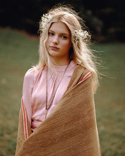 A young woman with blonde hair wearing a floral crown and pink top stands in a green field with a brown blanket draped over her shoulders.