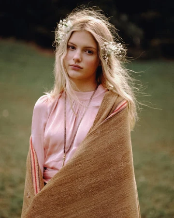 A young woman with blonde hair wearing a floral crown and pink top stands in a green field with a brown blanket draped over her shoulders.