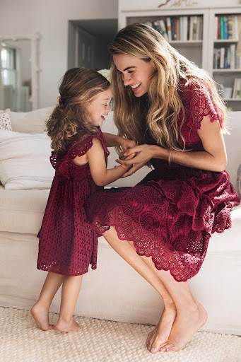A mother and young daughter share a joyful moment indoors, both wearing matching dark red lace dresses while sitting on a white sofa.