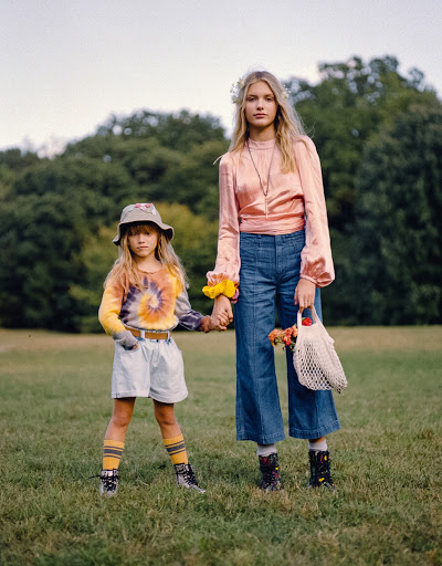 Two girls in vintage-style clothing stand holding hands in a grassy field with trees in the background in a fashion editorial shot.