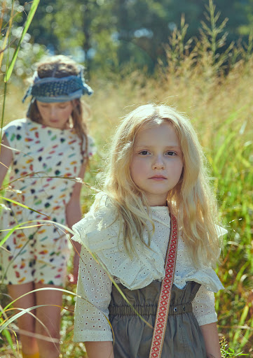 Two young girls in a field of tall grass, one with blond hair wearing a white eyelet top, the other with a blue headband. 
