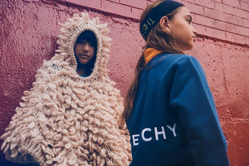 Two children pose against a red brick wall, one in a large knitted cape with a hood and the other in a blue Givenchy sweatshirt.