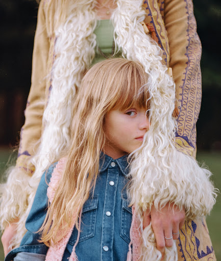 Young girl with blonde hair in a denim shirt and pink vest, posing with an adult with patterned coat in outdoor setting.