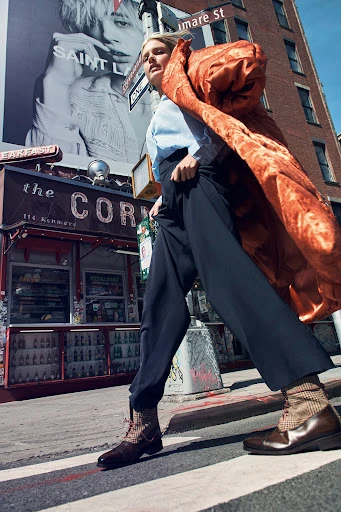 A stylish adult woman walks across a street in an urban environment. She wears a blue top, black pants, brown shoes and an orange shawl.