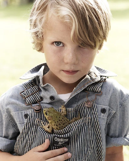 Young blonde boy wearing OshKosh overalls holding a green frog in his pocket, outdoors, medium shot, child fashion, close-up.