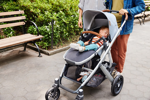 Young child sleeping peacefully in a stroller, with adults nearby in an outdoor park setting with benches and greenery.
