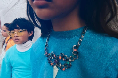 Close-up of child wearing a blue sweater and statement necklace, with other children visible in background.