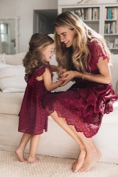 Smiling mother and young daughter wearing matching burgundy lace dresses while seated together on a sofa in a living room