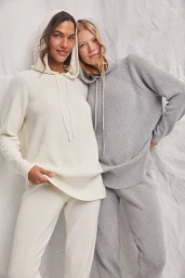Two female models in knit hooded sweaters and matching pants posing against a textured white backdrop.