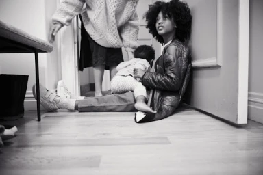 Black and white editorial portrait of a young woman seated on a wooden floor leaning against a doorway with a child.