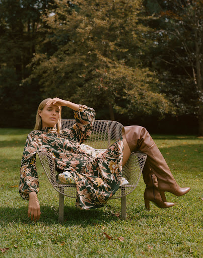 Woman in floral dress sits in metal chair on green lawn in outdoor fashion shot with trees, natural light, and over-the-knee boots.
