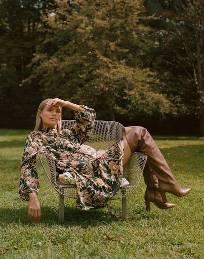 Woman in floral dress sits in metal chair on green lawn in outdoor fashion shot with trees, natural light, and over-the-knee boots.