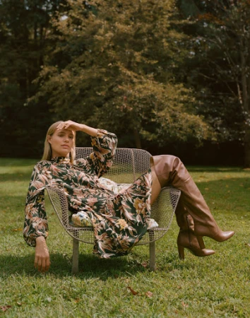 Woman in floral dress sits in metal chair on green lawn in outdoor fashion shot with trees, natural light, and over-the-knee boots.
