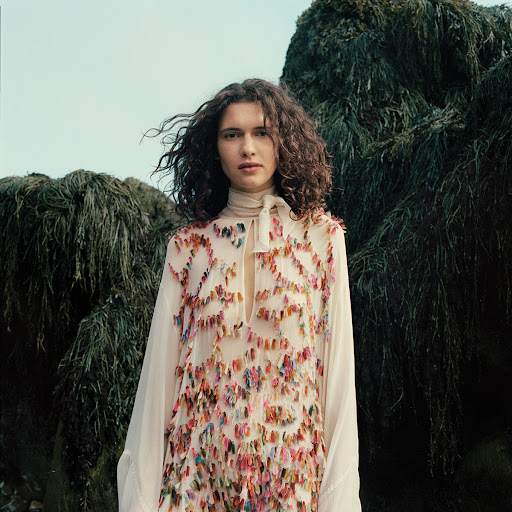 Adult female with curly hair wearing a flowing white dress with colorful embellishments poses outdoors near a mossy rock formation.