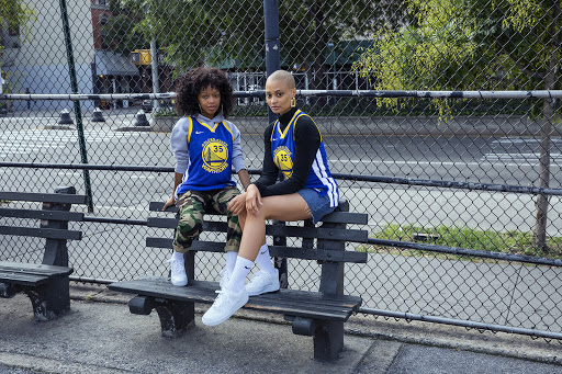 Two women in basketball jerseys sit on a park bench, showcasing urban style in daylight with trees and buildings in the background.