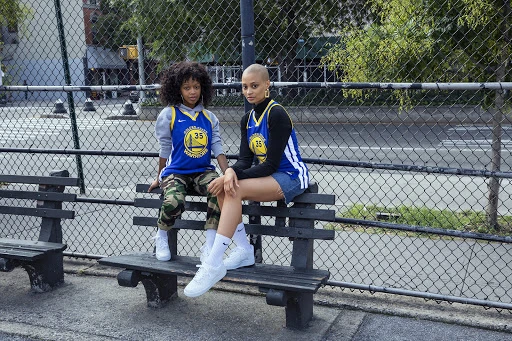 Two women in basketball jerseys sit on a park bench, showcasing urban style in daylight with trees and buildings in the background.