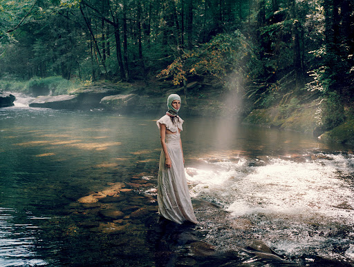A fair-skinned woman stands waist-deep in a river surrounded by a green forest. Sunlight streams through the trees onto the water.