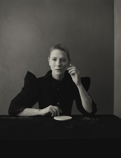 Portrait of Cate Blanchett wearing a black structured top with statement shoulders, seated at a dark table against a moody studio wall