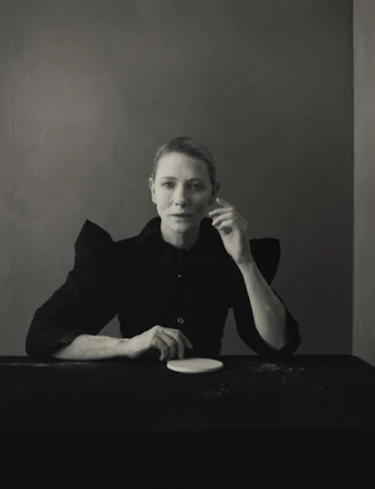 Portrait of Cate Blanchett wearing a black structured top with statement shoulders, seated at a dark table against a moody studio wall