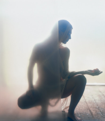 A nude woman crouches in front of a frosted glass panel, holding a small red sphere in her open palm, creating a soft, ethereal effect.