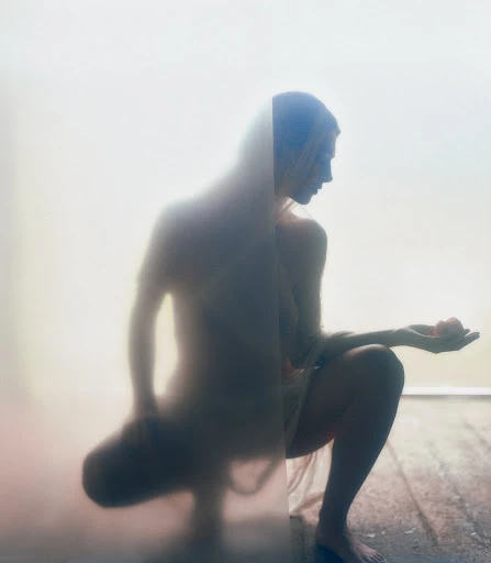 A nude woman crouches in front of a frosted glass panel, holding a small red sphere in her open palm, creating a soft, ethereal effect.