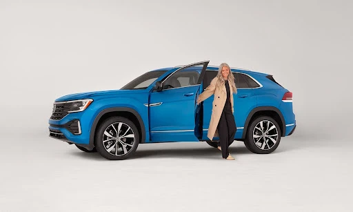 Blue Volkswagen Atlas Cross Sport SUV with a woman in a tan trench coat stepping out on a white studio backdrop for automotive advertising.