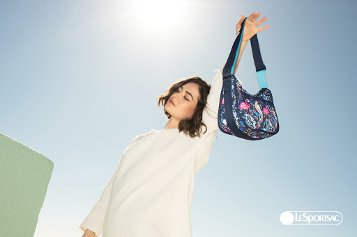 Brunette woman in a white dress holds up a patterned navy blue LeSportsac hobo bag against a bright blue sky in a sunny outdoor setting.