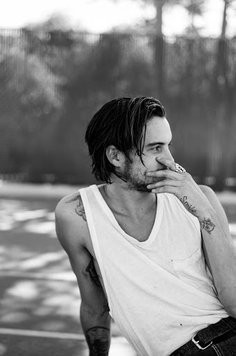 Monochrome portrait of a young adult man with dark hair, wearing a white tank top and jeans, resting his hand on his face.