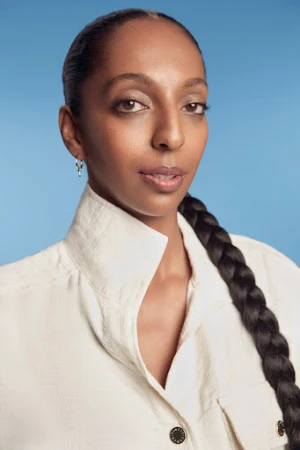 Portrait of a woman wearing a cream colored button down shirt with a high collar against a blue background