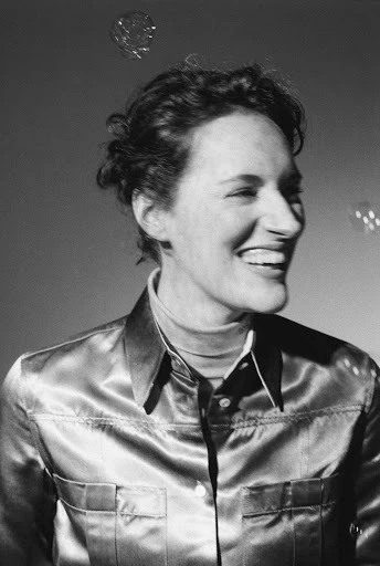 Portrait of a smiling woman in a shiny collared shirt, close-up shot, against a gradient grey background with bubbles floating in the air.