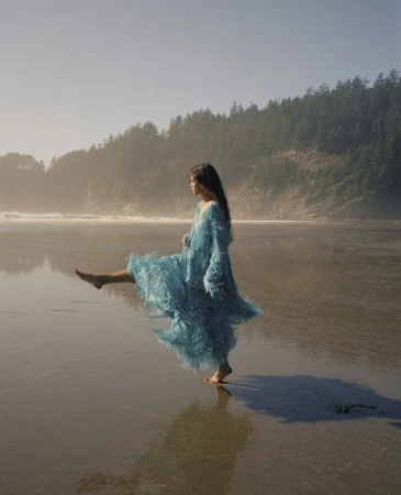 Woman in a textured blue fringed dress kicking up water on a sandy beach under hazy sunlight with distant trees.
