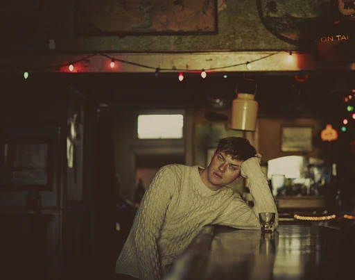 Young man in a cream cable knit sweater leaning on a wooden bar in a dimly lit tavern with string lights.