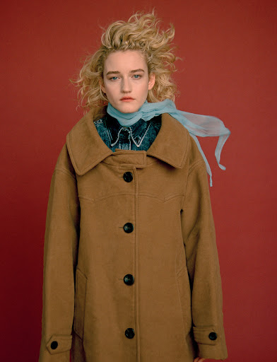 Portrait of a blonde woman with a tan coat and blue scarf standing in front of a solid red background in a fashion editorial style.