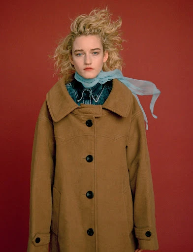 Portrait of a blonde woman with a tan coat and blue scarf standing in front of a solid red background in a fashion editorial style.