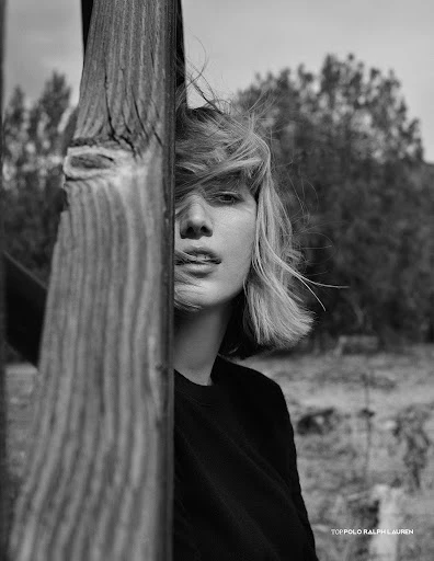 Monochrome outdoor portrait of a blonde woman with windblown hair, standing next to a wooden structure, against a blurred landscape.