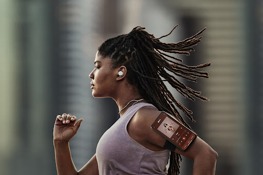 A woman with locs runs outside, wearing wireless earbuds and an armband phone holder. She is wearing a mauve tank top.