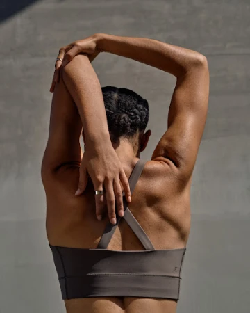 Woman wearing a brown sports bra stretching arms overhead against a grey wall