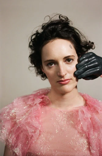 Portrait of a woman with short curly hair wearing a pink tulle top, with a black-gloved hand touching her face.