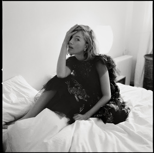 Monochrome portrait of a woman kneeling on a white bed wearing a dark patterned dress with her hand to her forehead