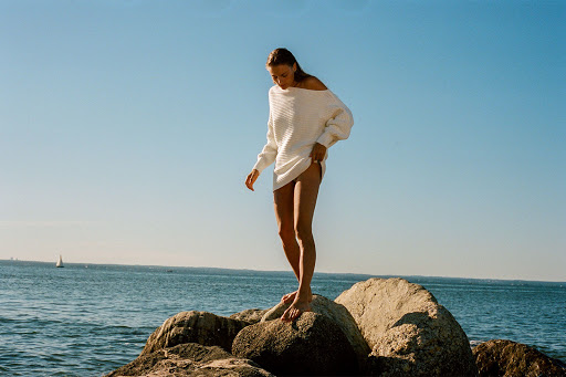 Woman in an off-the-shoulder white knitted sweater standing barefoot on coastal rocks above ocean water.