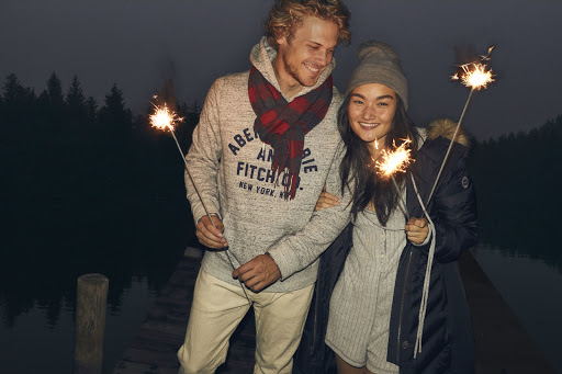 Young couple smiles on a dock at dusk, holding sparklers, wearing casual jackets and beanies against a dark lake and forest backdrop.